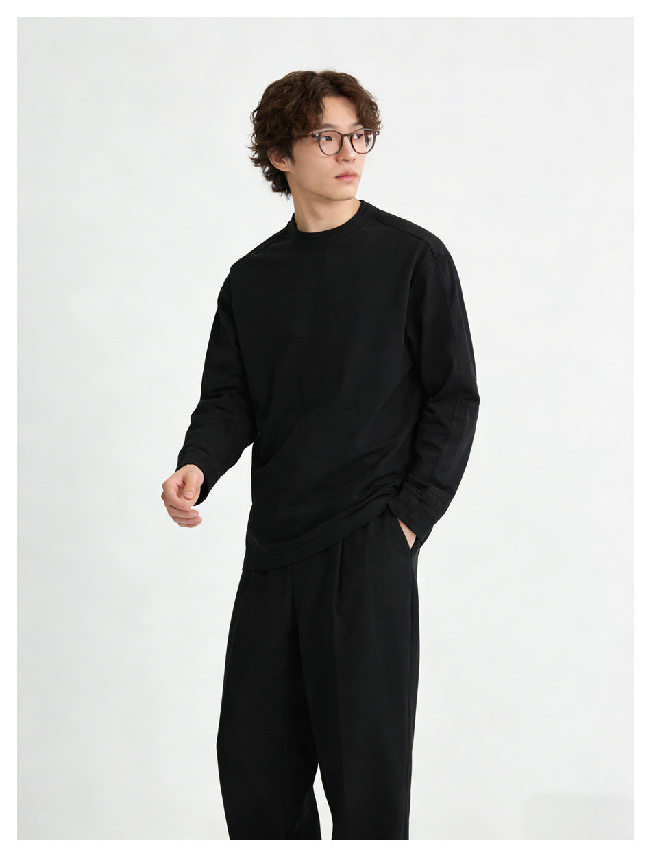 Man wearing black long-sleeve crew-neck top with black trousers, one hand in pocket