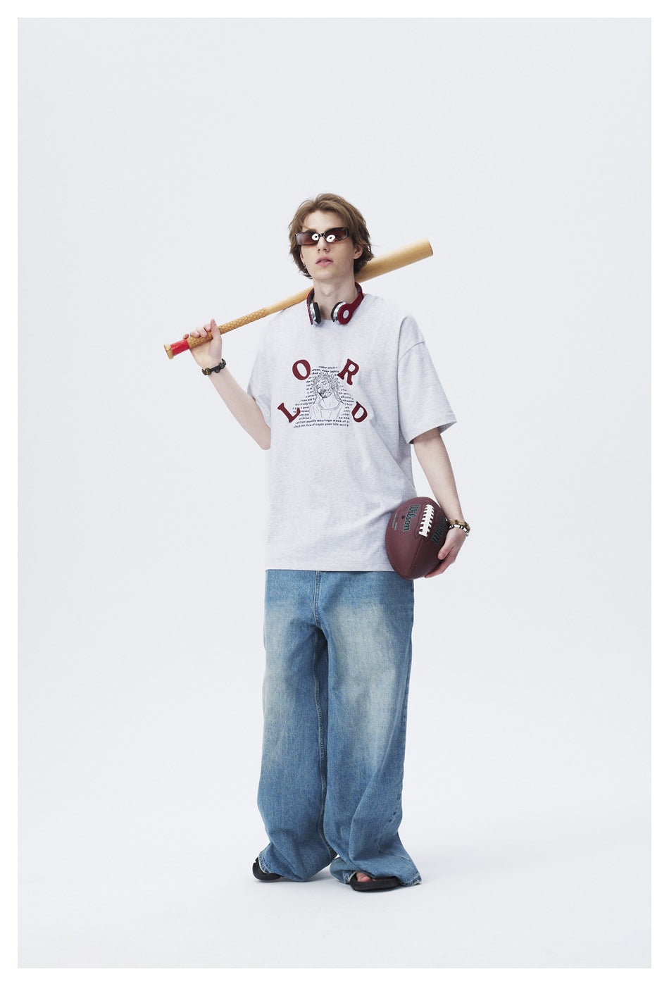 Model in light gray LORD graphic T-shirt and wide-leg jeans holding a baseball bat and football