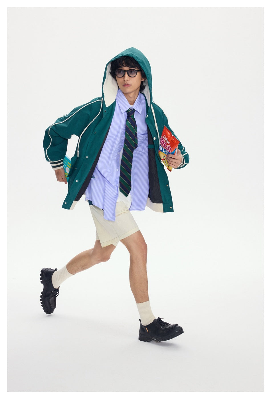 Model wearing a teal hooded jacket with cream piping over a striped shirt and tie, paired with beige shorts.
