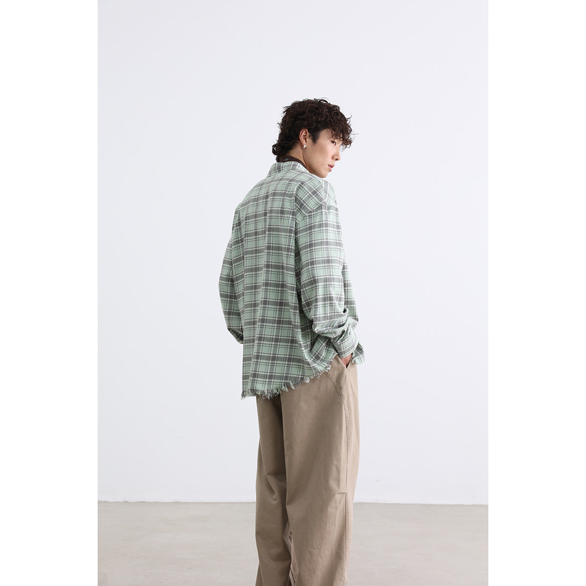 Side and back view of a model wearing a sage green plaid shirt with a frayed hem and tan trousers.
