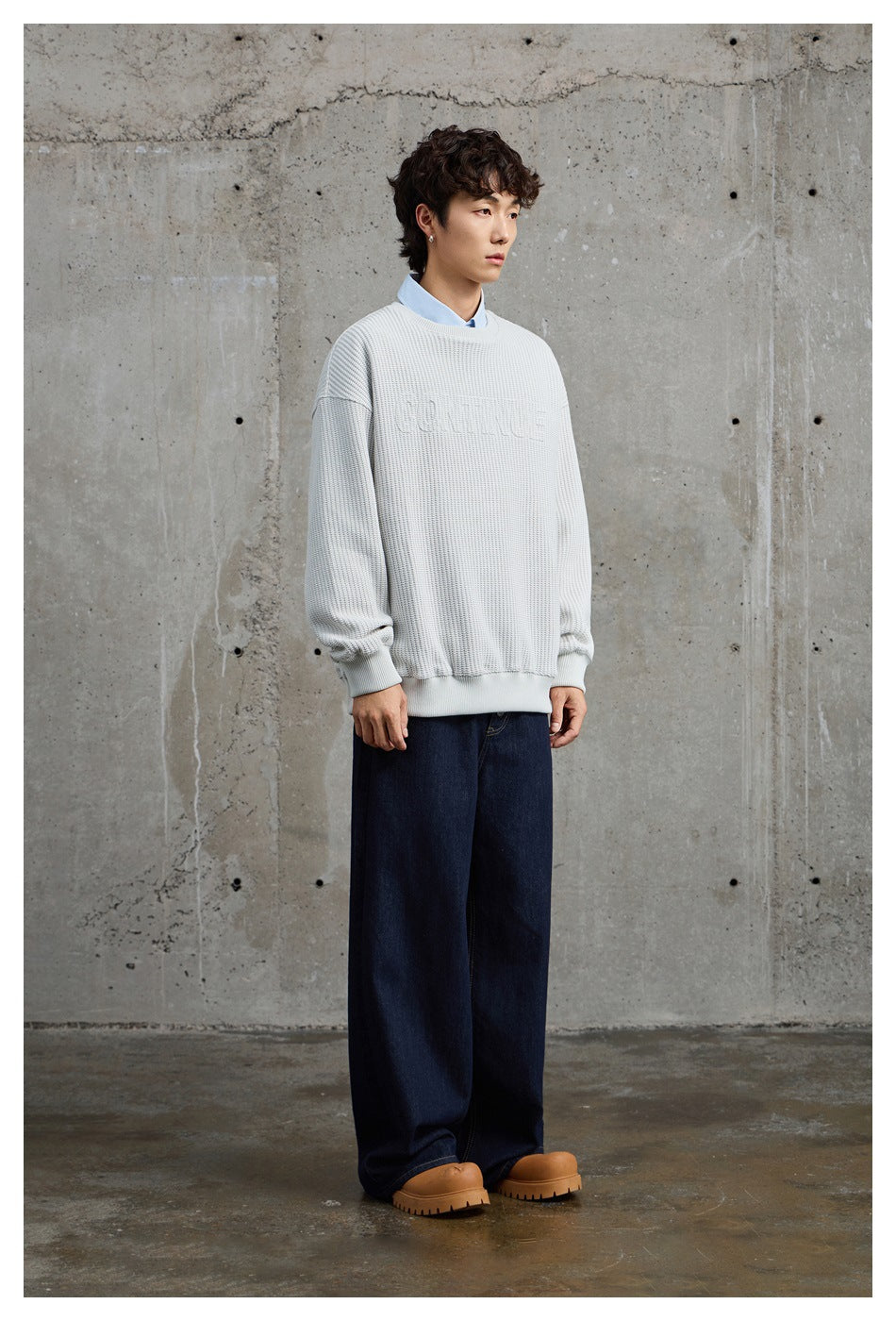 Full-length front view of a model in a light gray textured crewneck sweater over a pale blue shirt and dark wide-leg jeans.