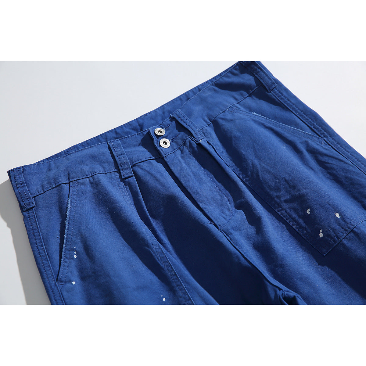 Close-up of cobalt-blue trousers showing the waistband, double metal buttons, front pocket and white paint-like speckles.