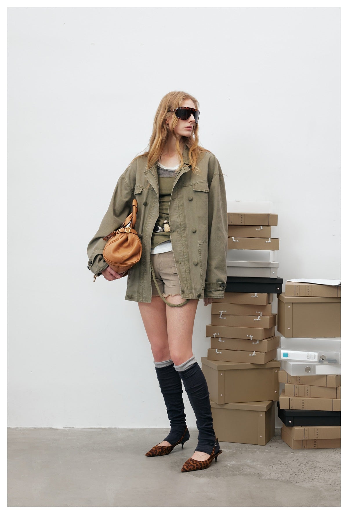 Woman in an open olive green oversized jacket over layered tops and shorts, angled view, standing by stacked boxes