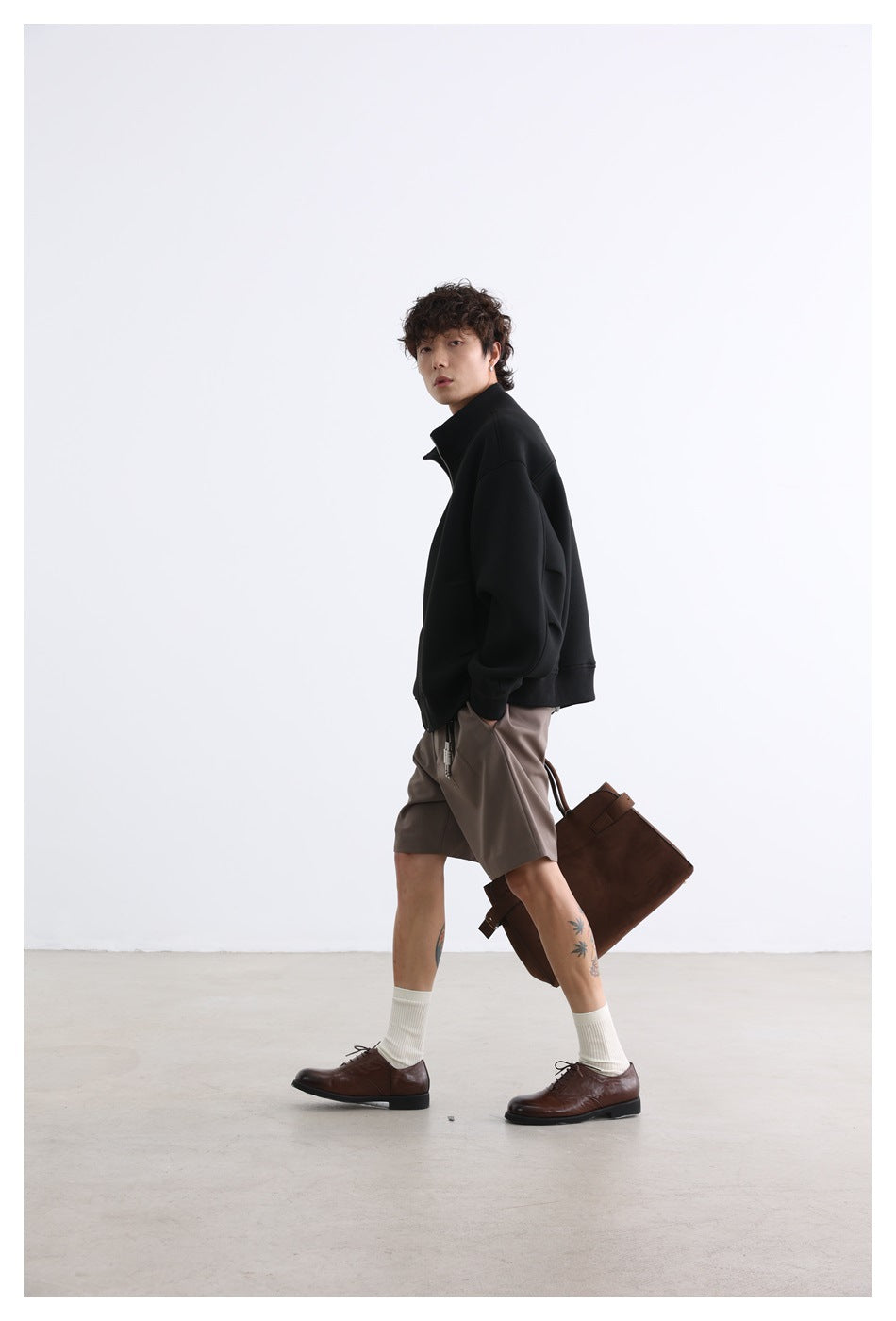 Model wearing a black stand-collar zip jacket and taupe shorts, shown walking in a side view.