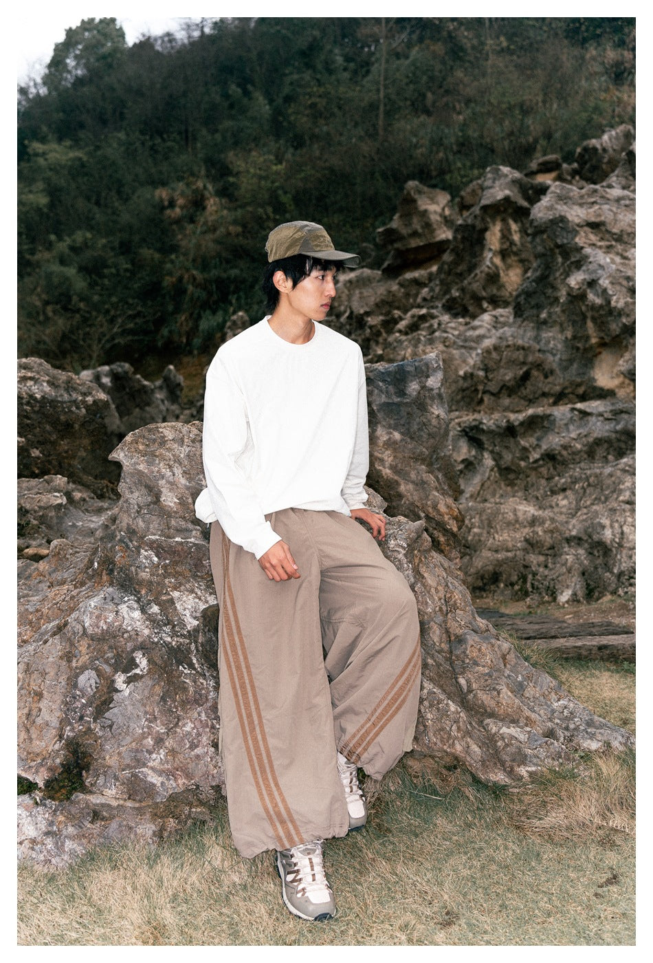 Model seated on rocks wearing a white long-sleeve top, taupe wide-leg pants with side stripes, and a cap.