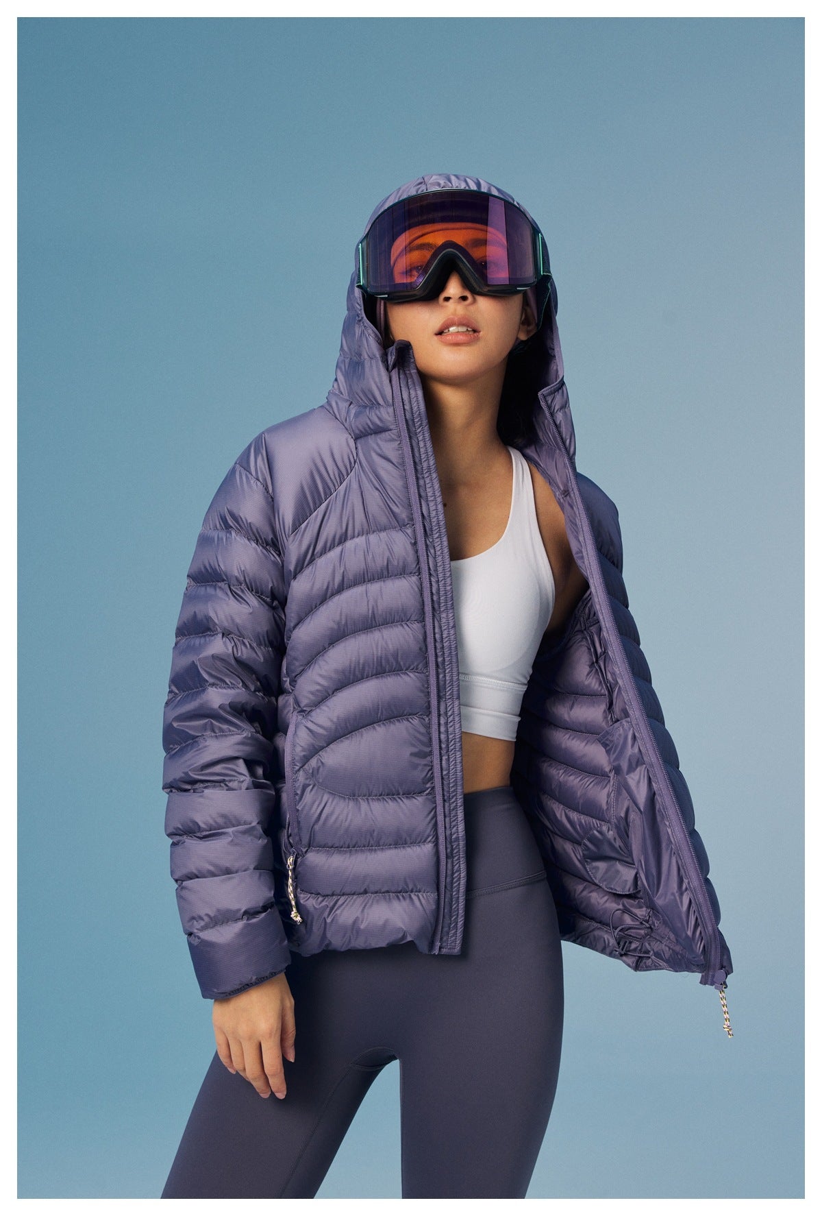 Woman wearing an open lavender quilted hooded puffer jacket, ski goggles, a white crop top and dark leggings.