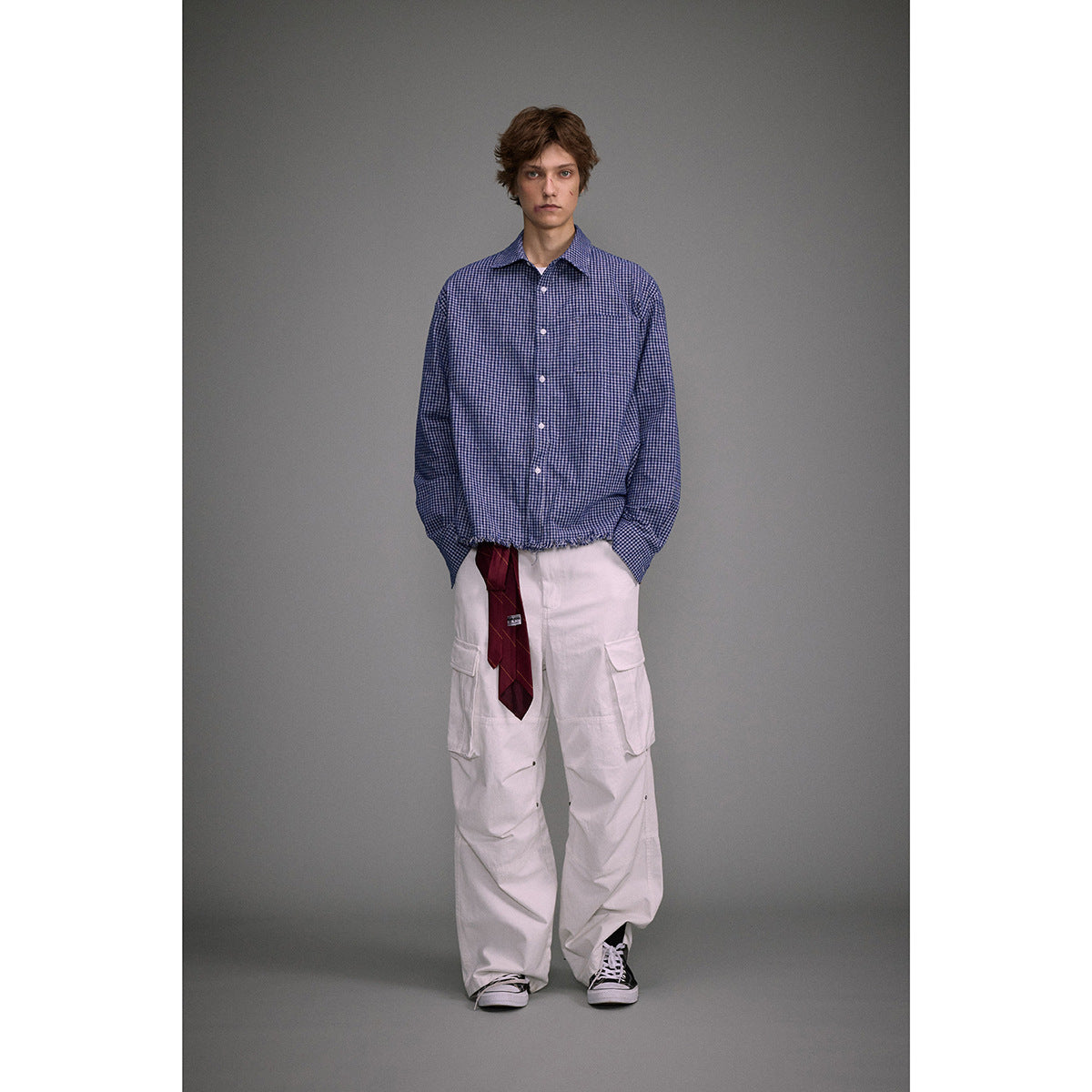 Model wearing a blue small-check button-up shirt with a gathered hem, white cargo pants, and a burgundy tie.