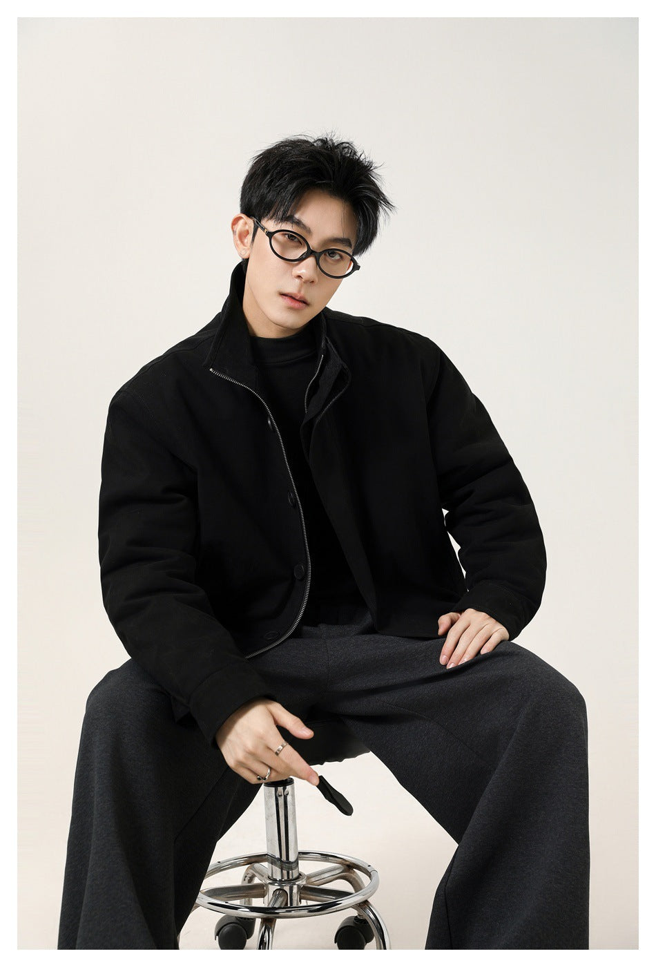 Seated model wearing a black high-collar jacket with a button-and-zip front and dark trousers.