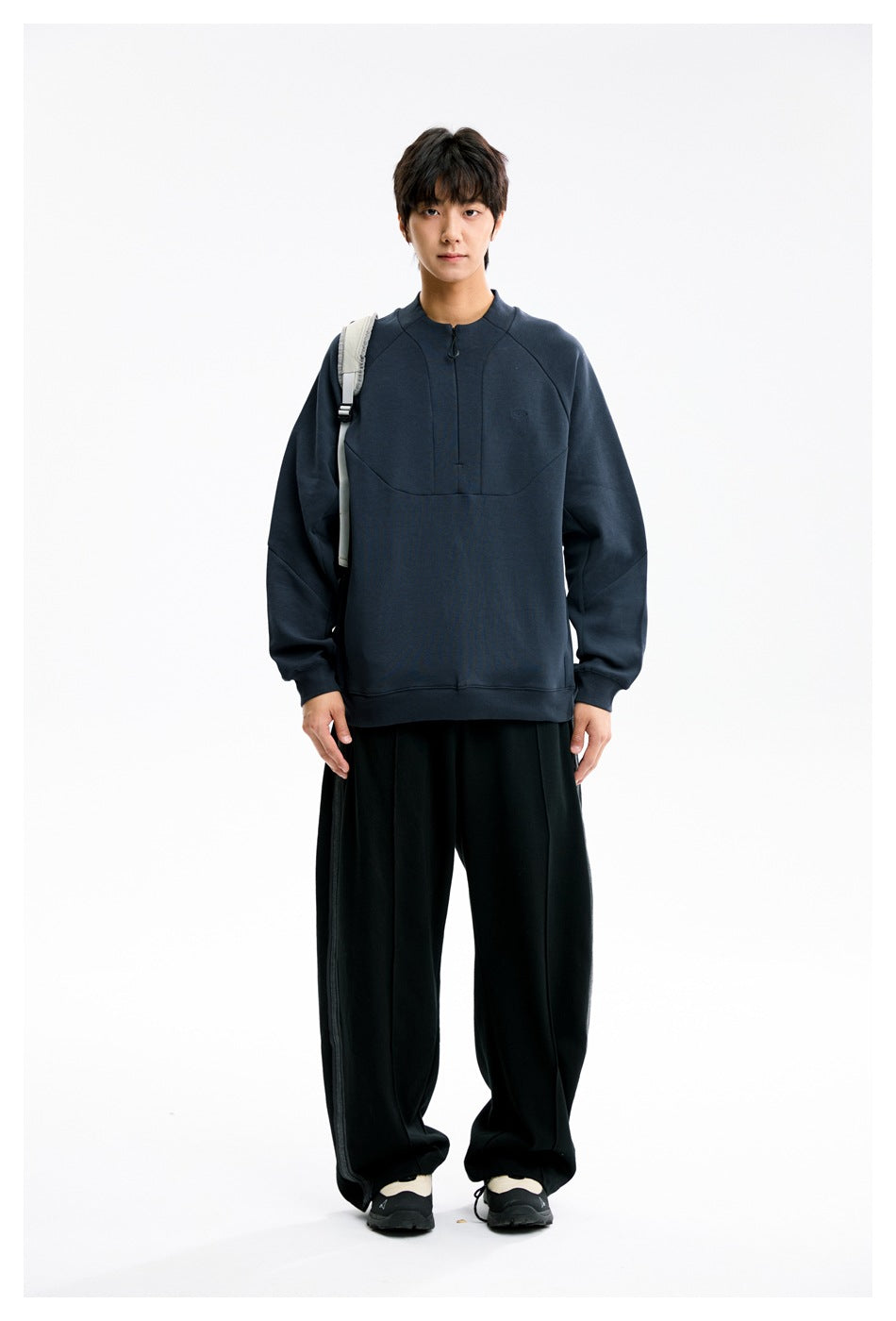 Model wearing a navy half-zip sweatshirt with black wide-leg pants, a gray backpack strap on one shoulder