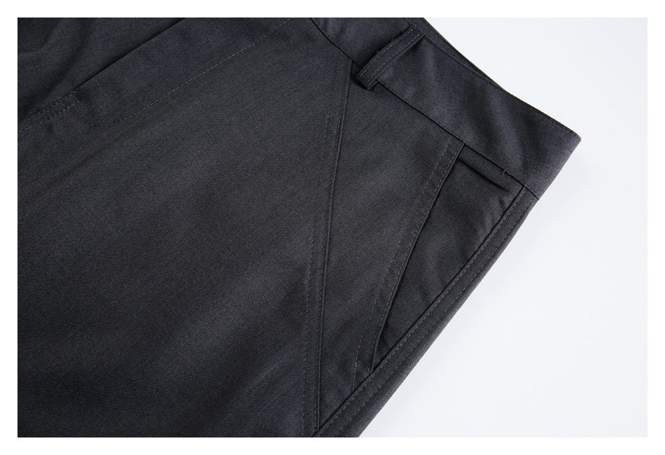 Close-up of charcoal trousers showing a slanted pocket opening, waistband, and stitched panel seams.