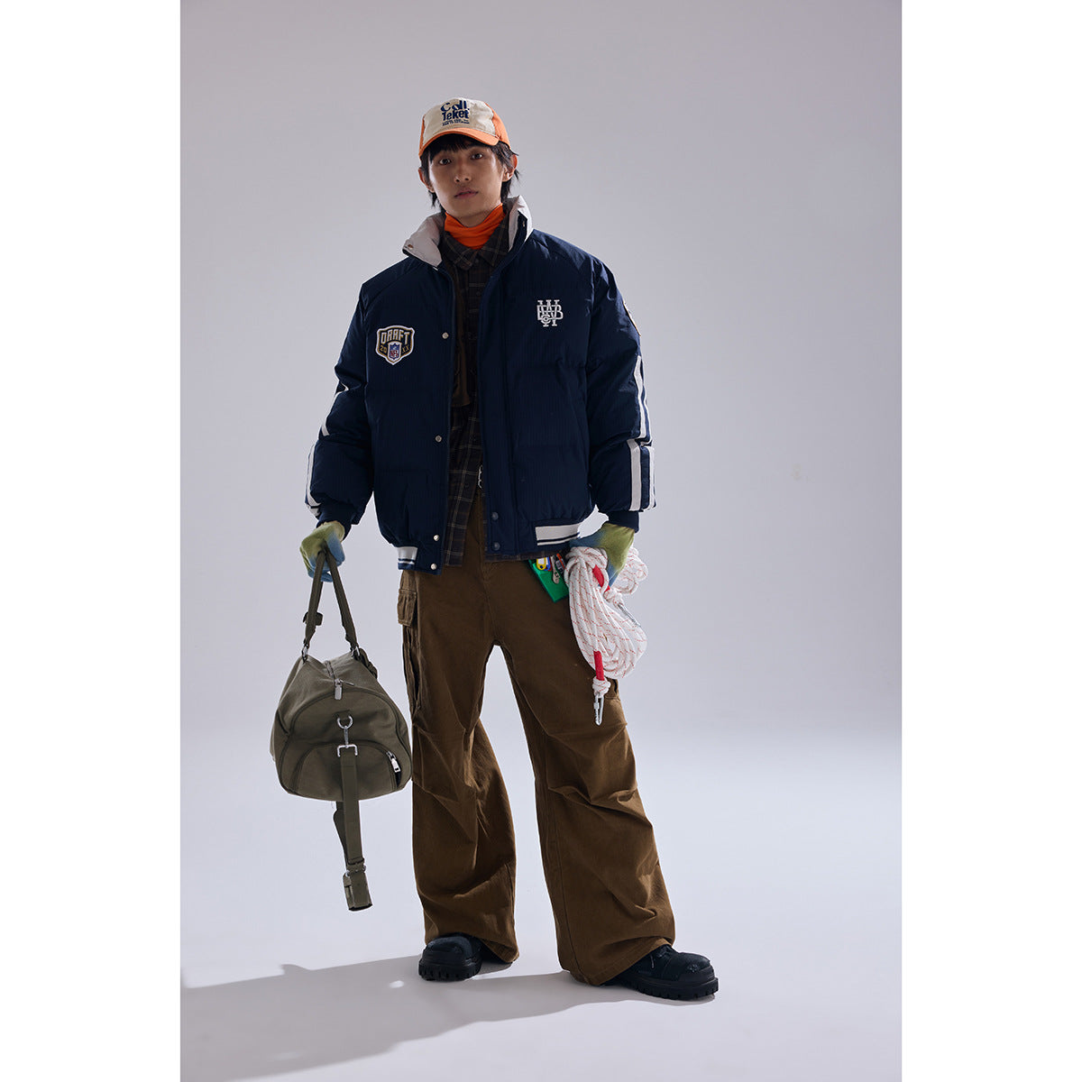 Front view of a model wearing a navy padded jacket with white stripes and patches, brown cargo pants, and an orange cap.