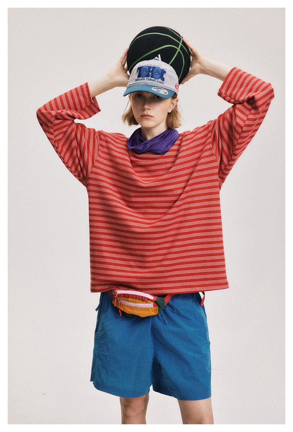 Front view of a model wearing a loose red-and-white striped shirt and blue shorts, holding a basketball overhead.