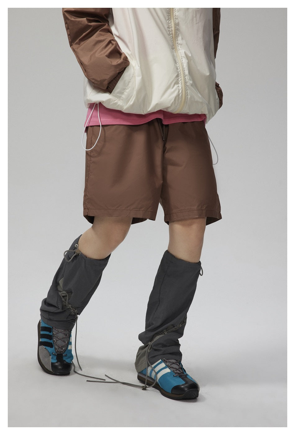 Angled front view of a person wearing a white jacket with brown sleeves, brown shorts, and dark gaiters over blue shoes.