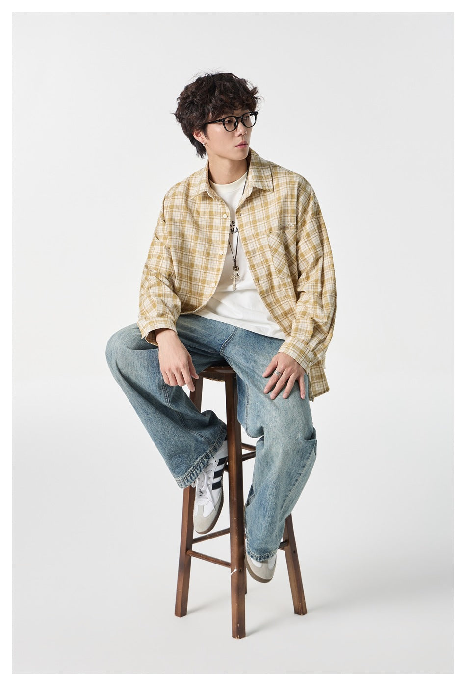 Model seated on a stool in a mustard and cream plaid button-up shirt, white tee, and loose blue jeans.