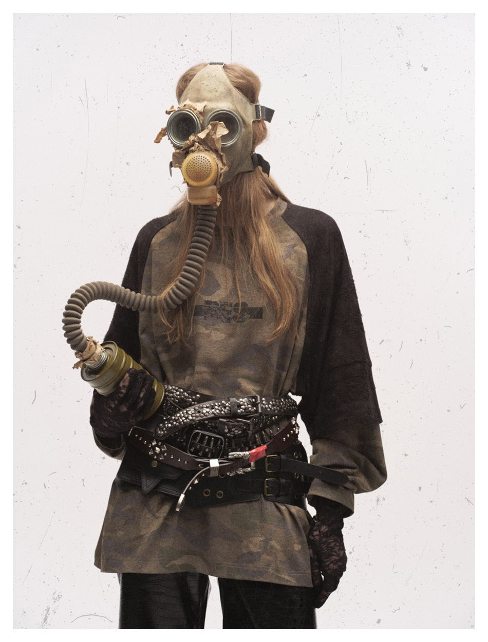 Standing model wearing an oversized brown camouflage raglan shirt with black sleeves and layered studded belts.
