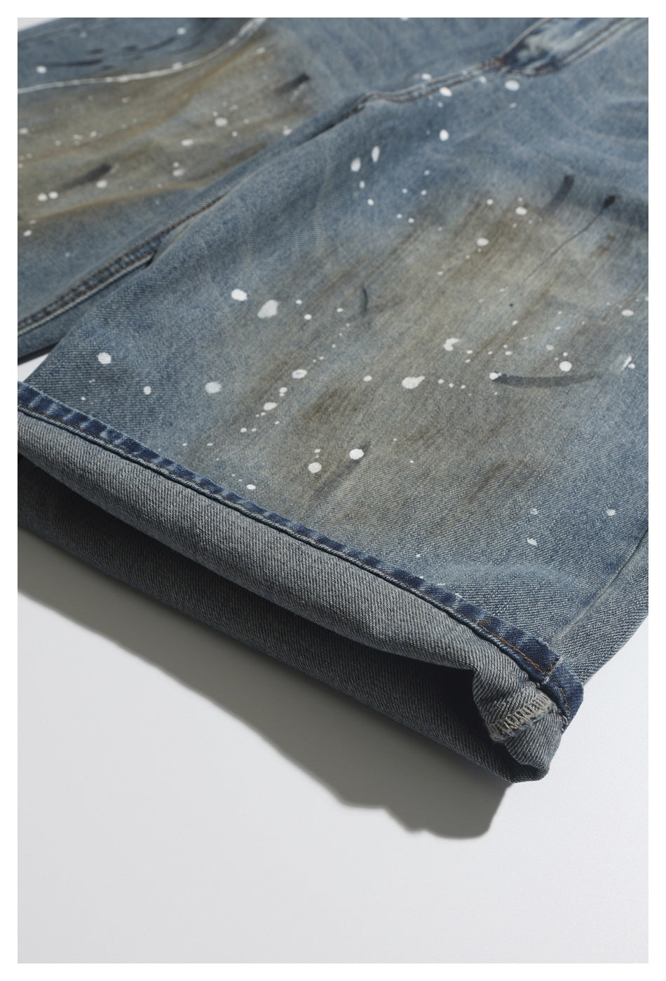 Close-up of a turned-up jeans cuff showing blue-gray denim, white paint splatters, and seam stitching.
