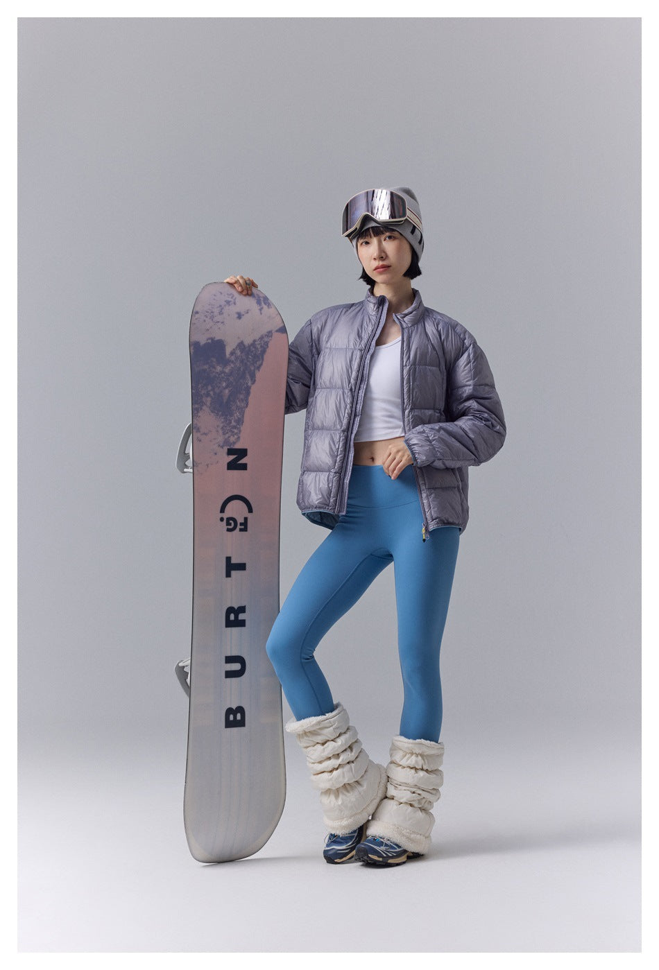 Model wearing a metallic gray quilted jacket with blue leggings, standing beside a snowboard.