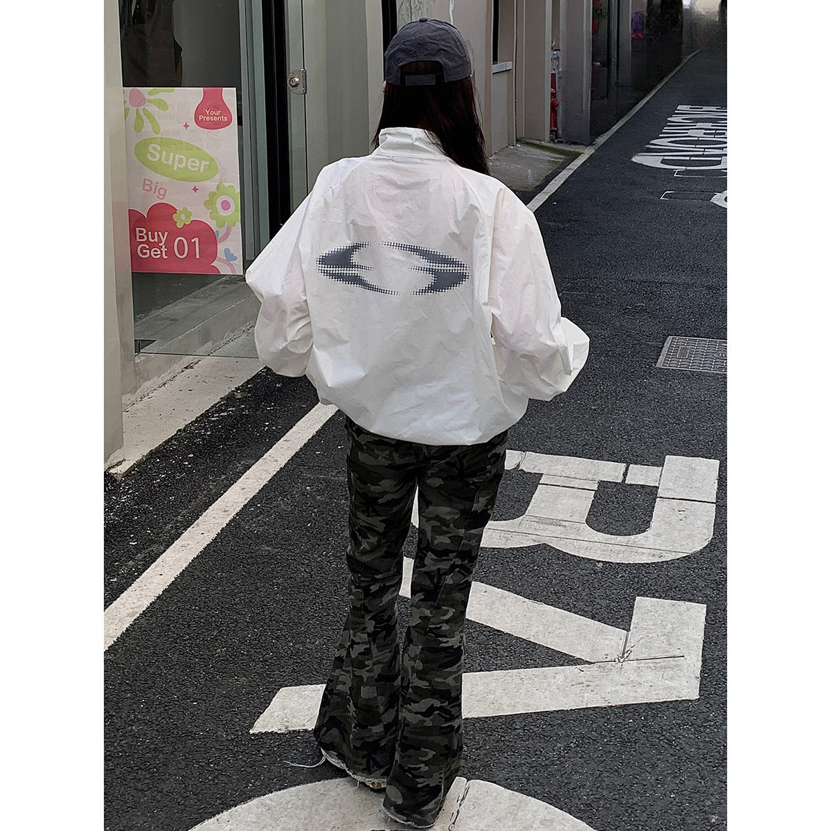 Back view of a model wearing a white jacket with a large dark emblem and dark camouflage flared pants.