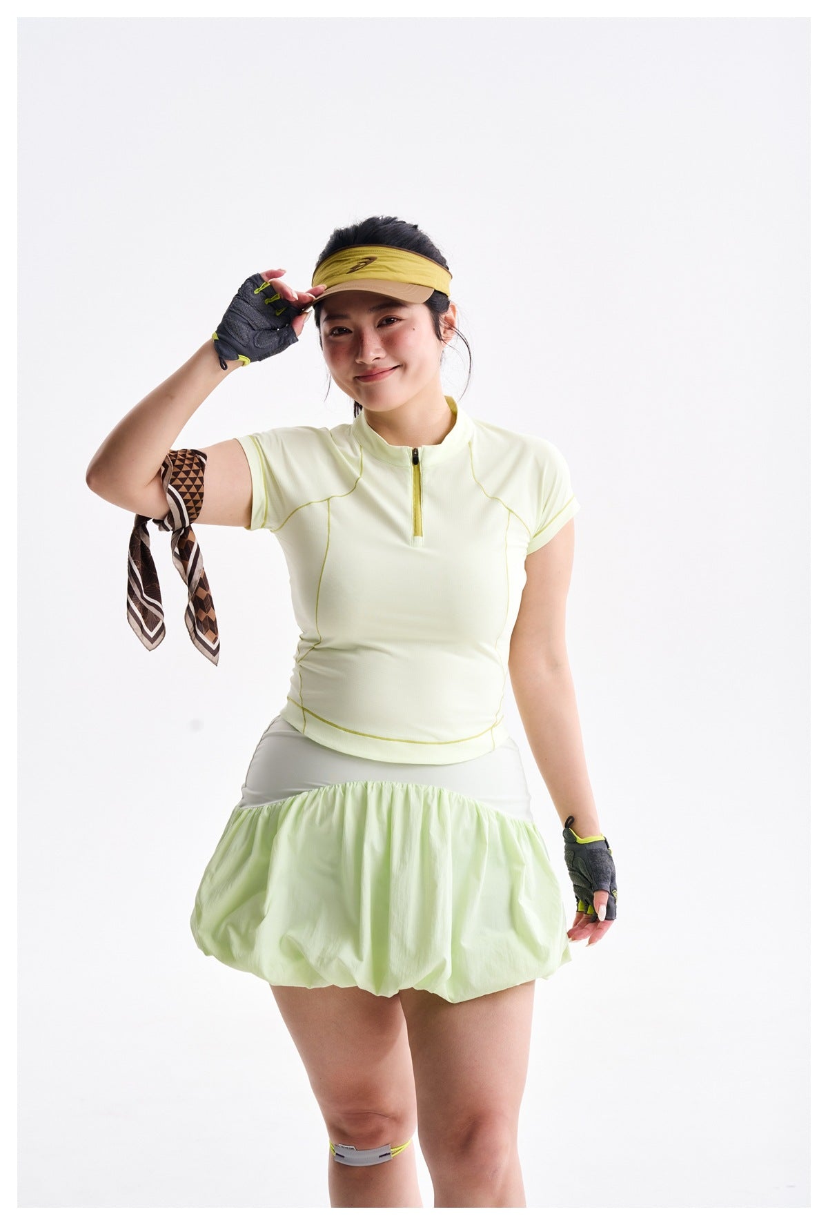 Model in a pale yellow-green zip-neck top and bubble skirt, shown from the waist up with gloves and a visor.