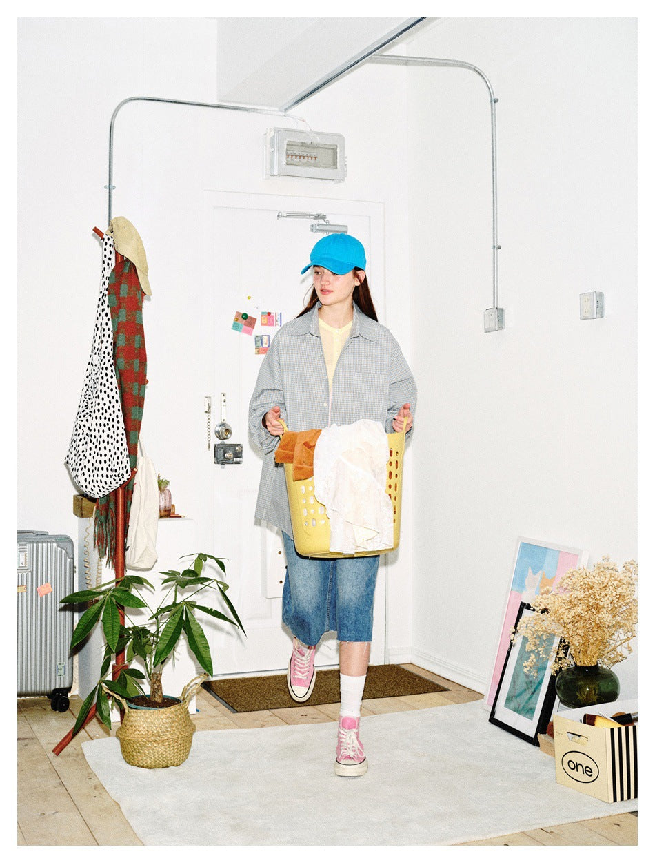 Model wearing a pale blue check button-up shirt, yellow top, denim shorts and blue cap, holding a laundry basket.