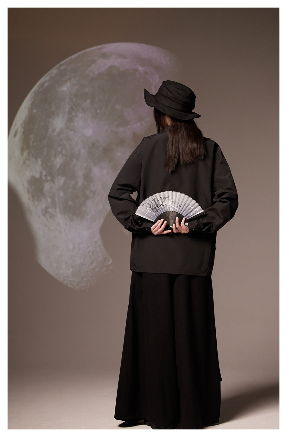 Rear view of a model in a black shirt and long skirt, holding an open fan behind her.