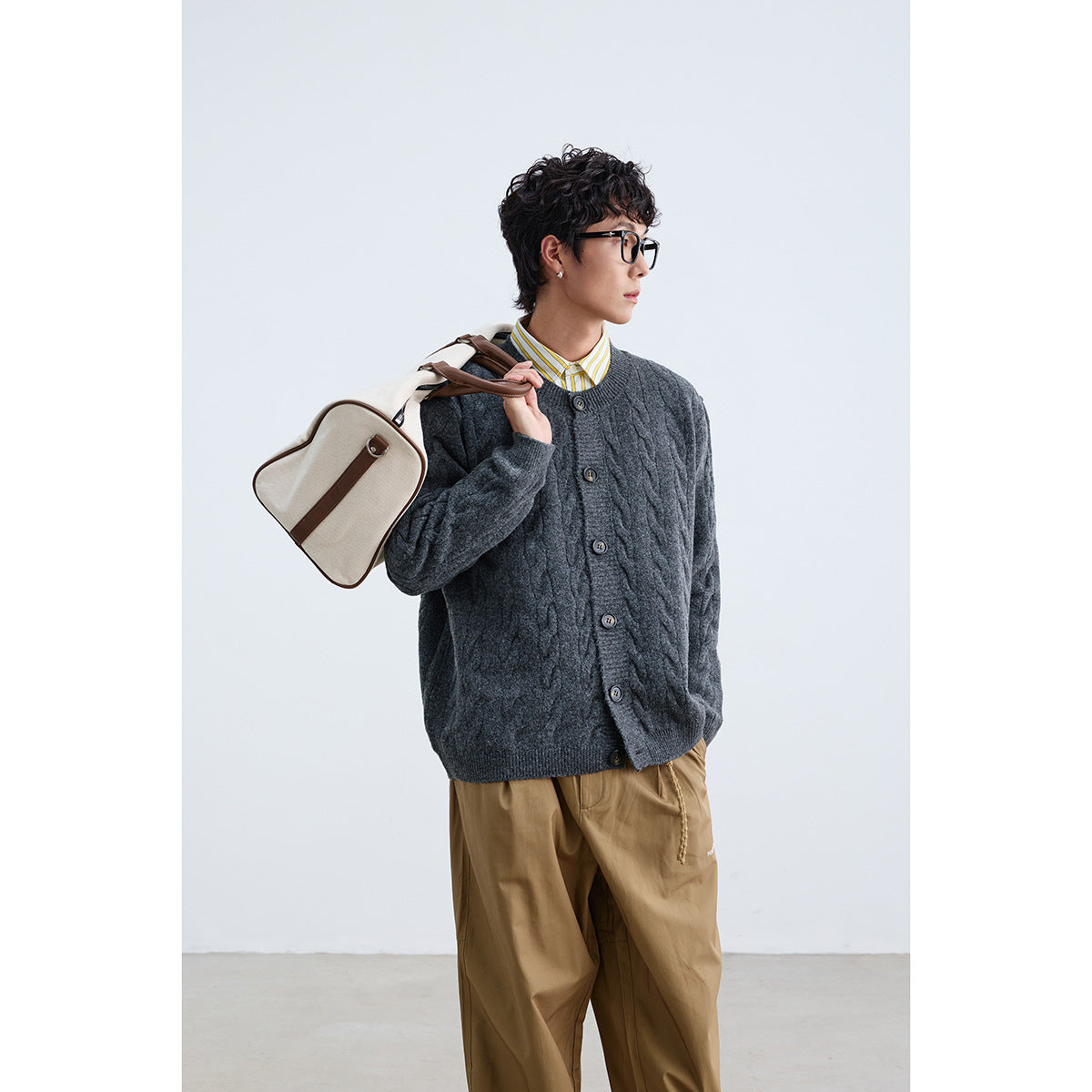 Model wearing a charcoal cable-knit button cardigan over a yellow-striped shirt with tan trousers.