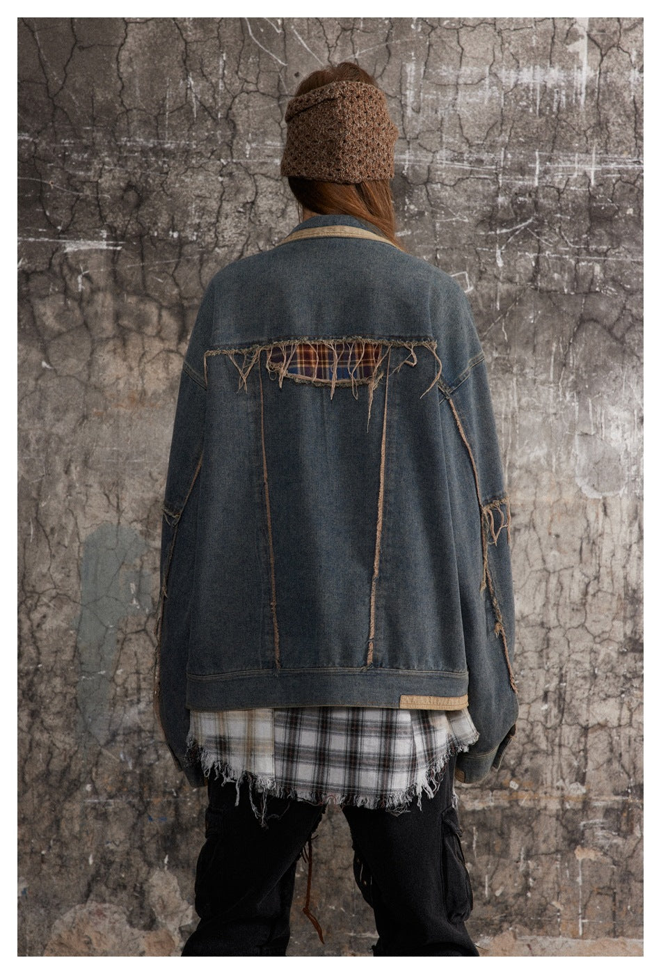 Back view of a model wearing an oversized distressed blue denim jacket with frayed seams and plaid accents.