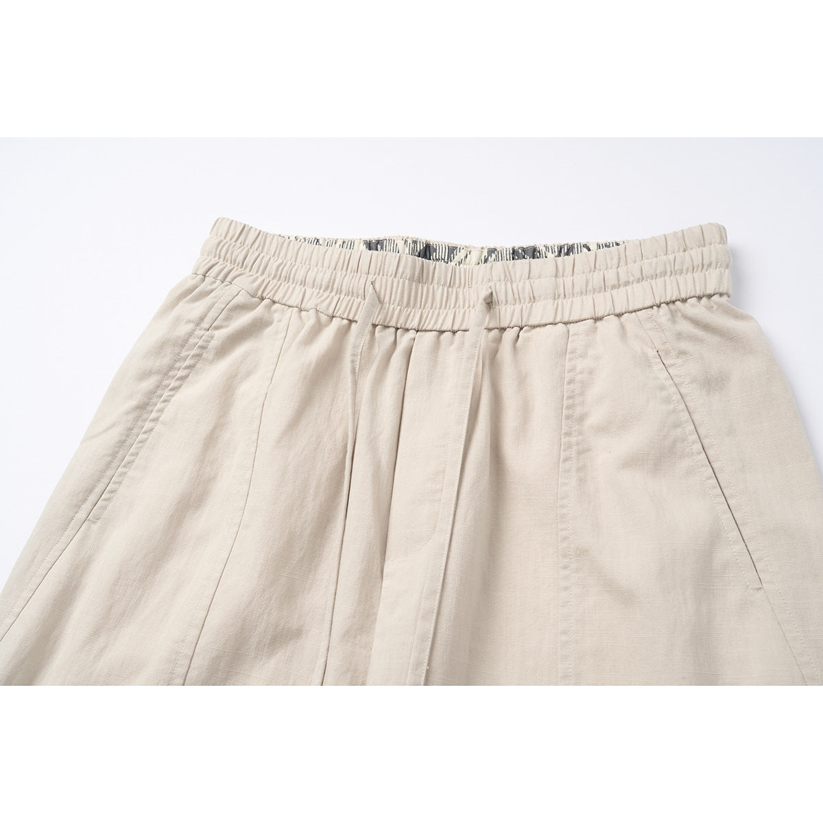 Close-up of light beige shorts showing the gathered elastic waistband, front pleats and angled pocket seams.