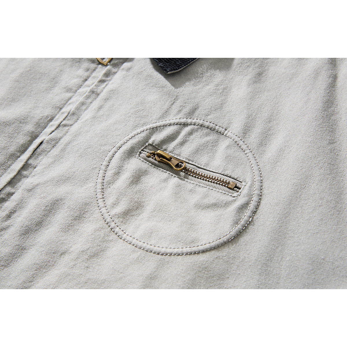 Close-up of a light gray jacket's round stitched chest pocket with a horizontal brass zipper.