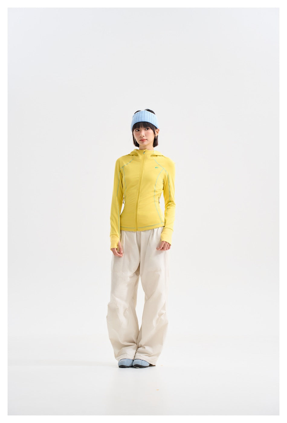 Model wearing a yellow zip-front hooded jacket with teal piping, loose cream pants, and a light blue headband.