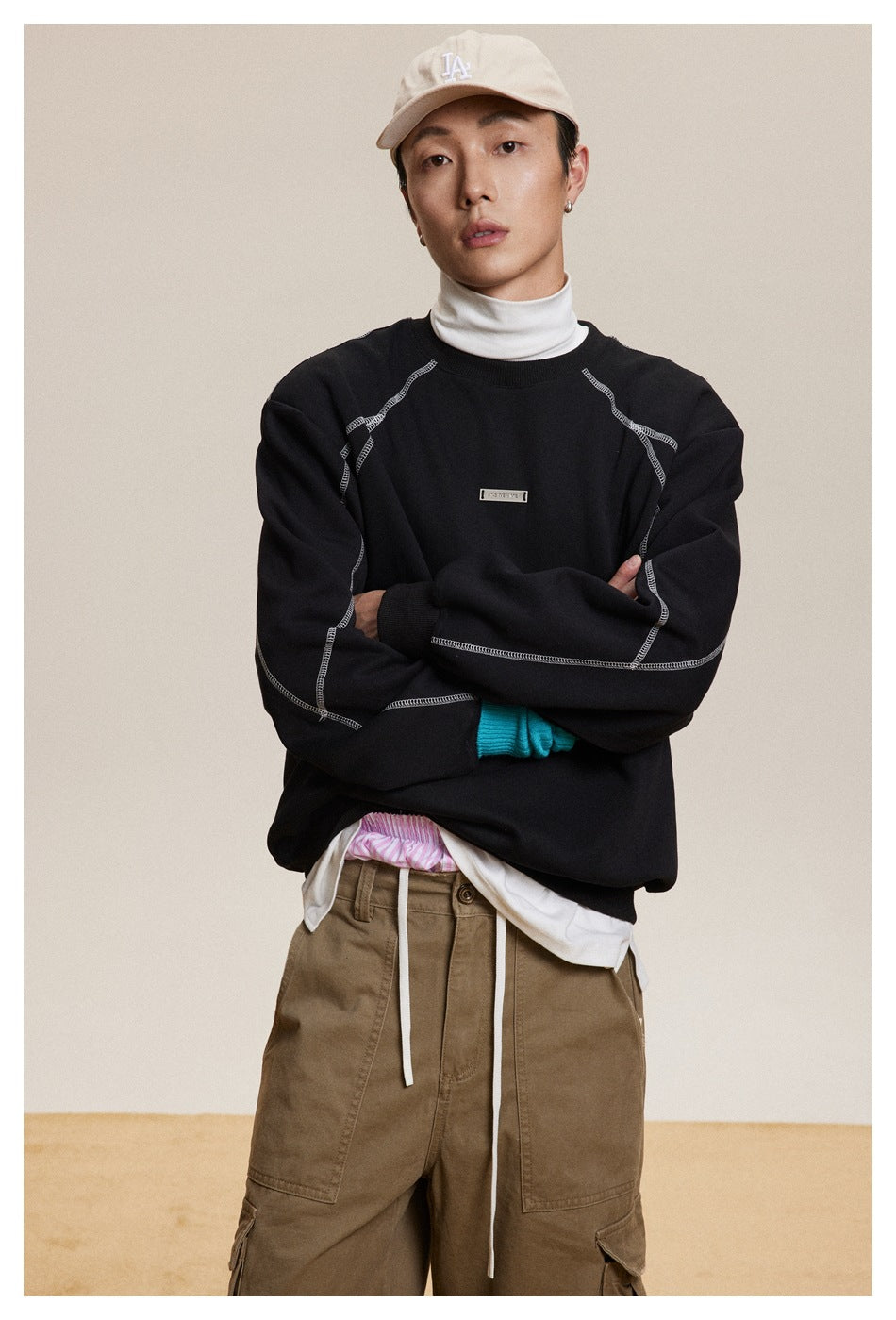 Model wearing a black crewneck sweatshirt with prominent white contrast seams and a small chest plaque.