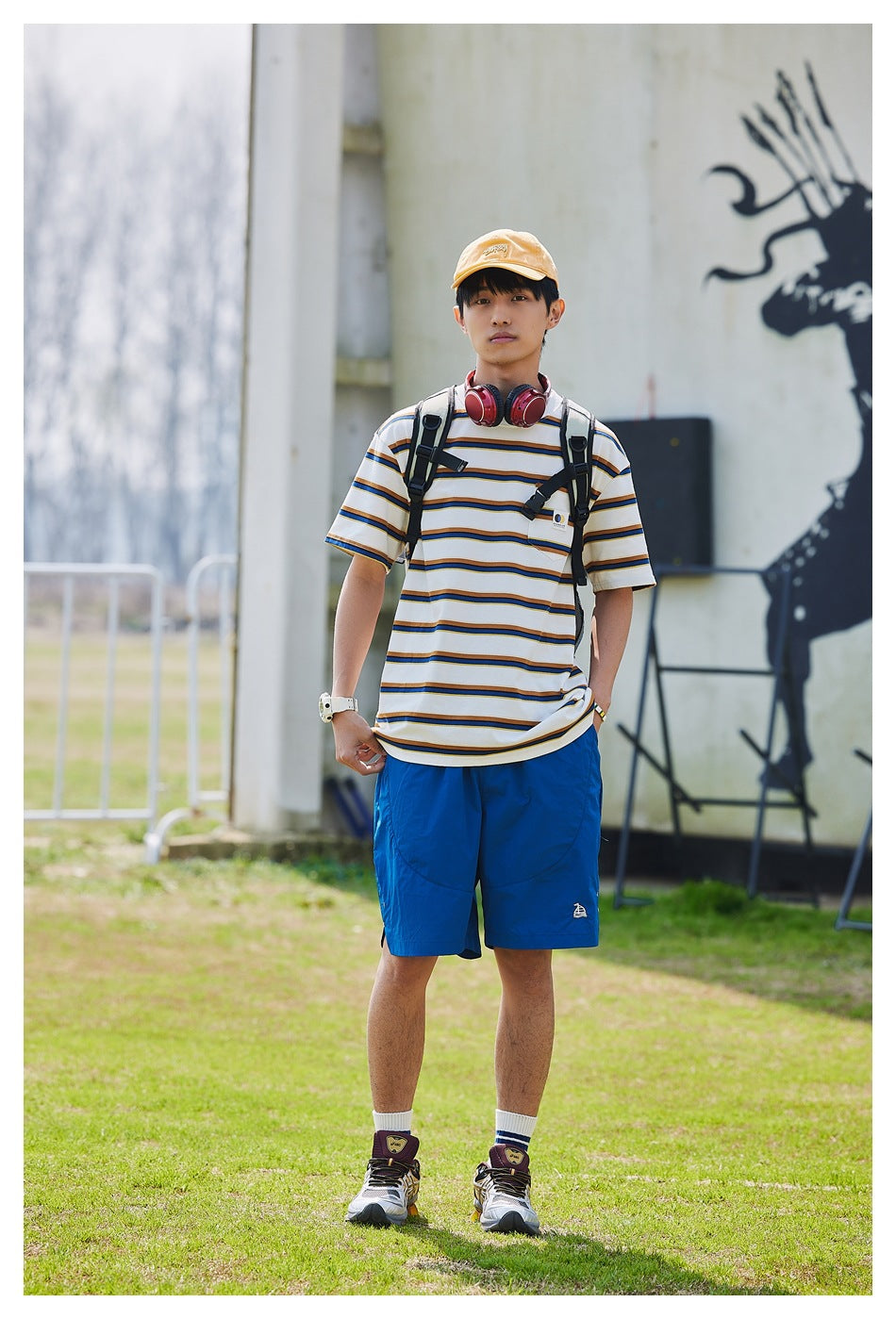 Model wearing the white striped pocket T-shirt with blue shorts, orange cap and backpack standing on grass