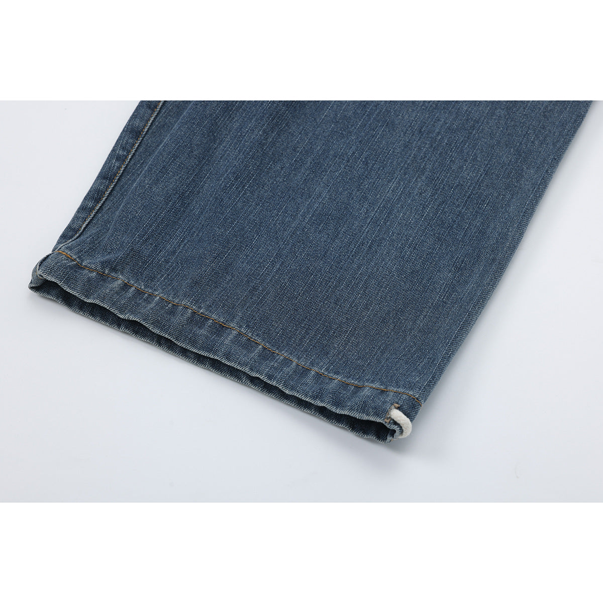 Close-up of a blue denim pant leg hem with tan stitching and a cream drawcord loop.