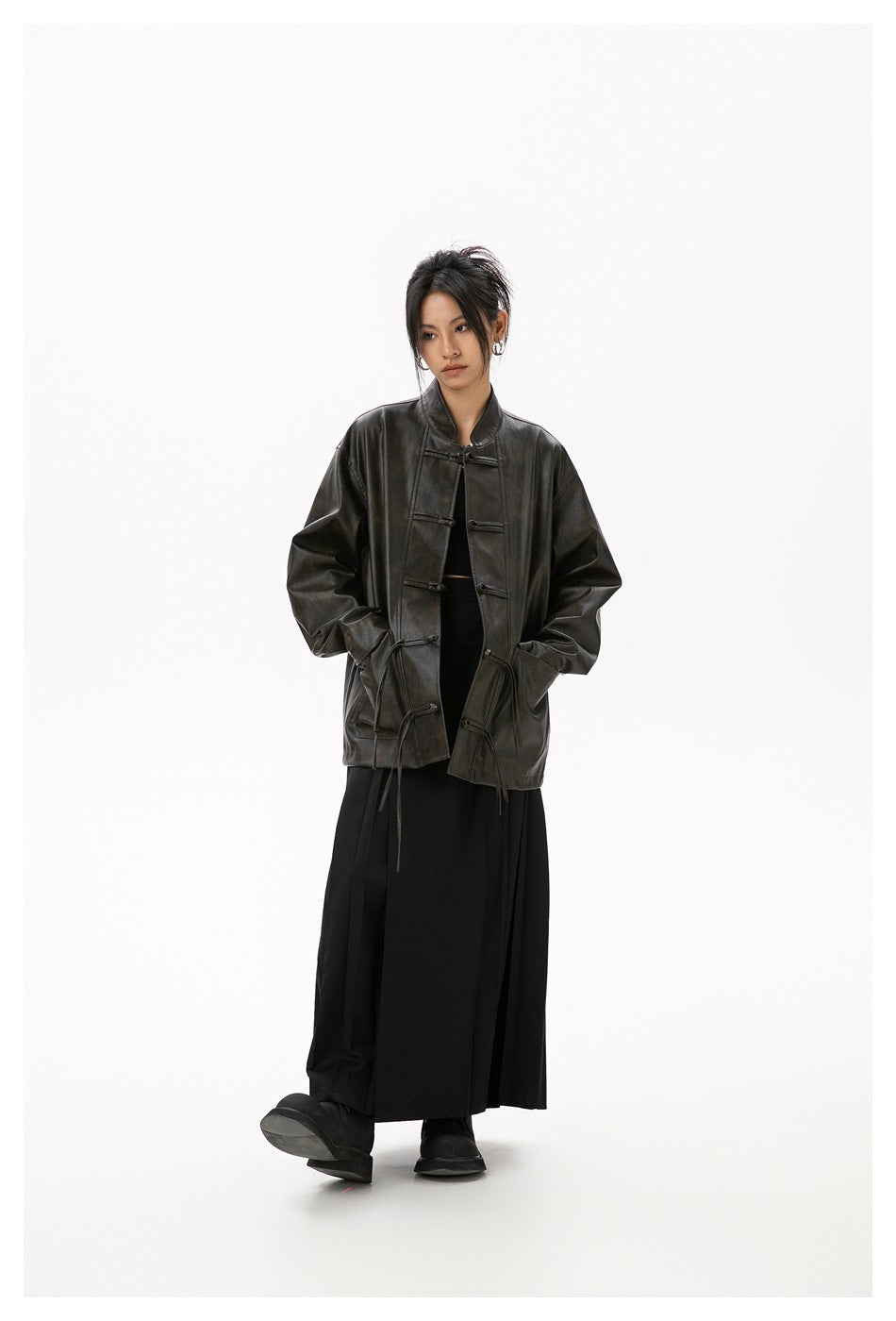 Model wearing the dark brown leather-look frog-button jacket open over black top and long black pleated skirt, full length