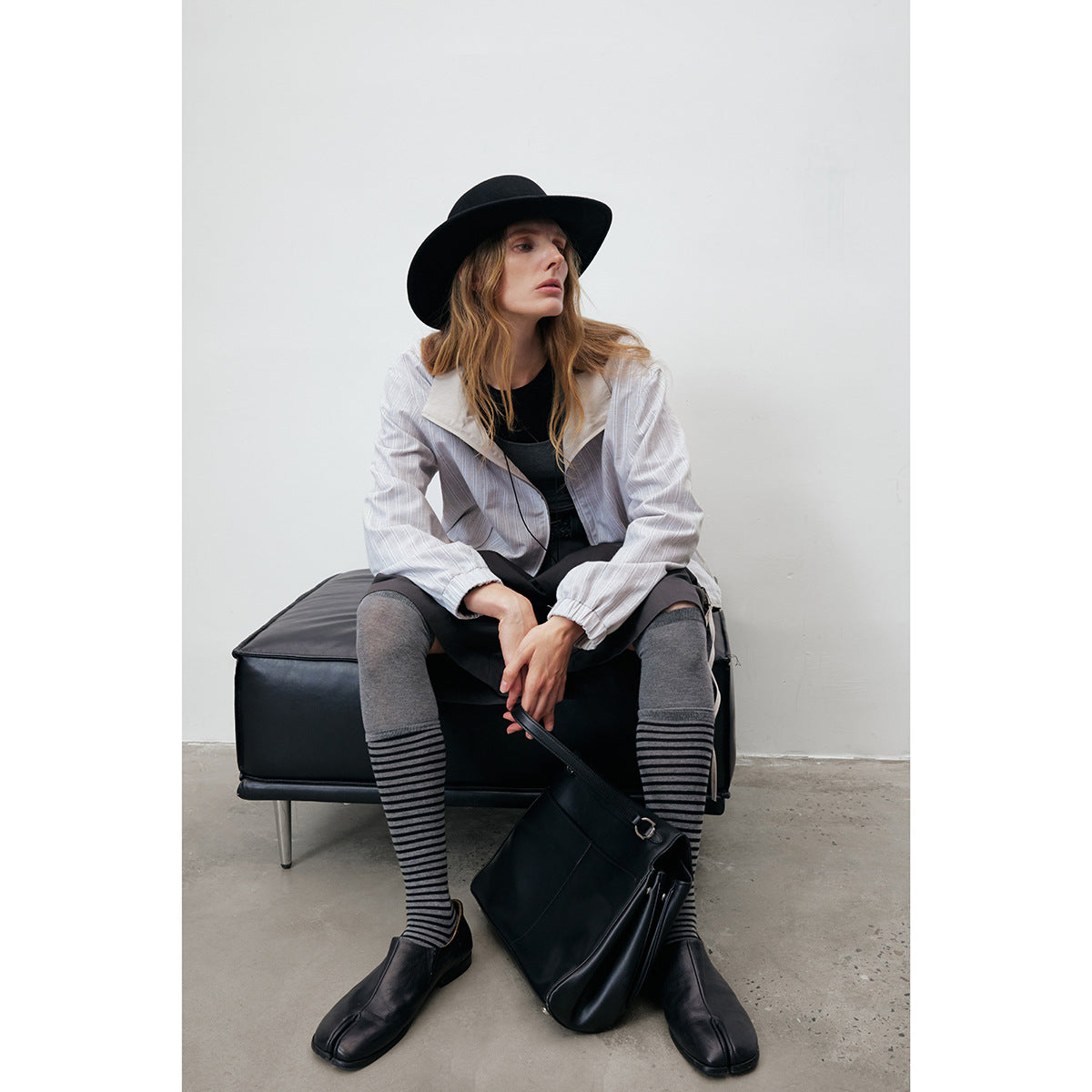 Model seated in a light gray pinstriped zip jacket, black hat, dark shorts, striped socks and black shoes.