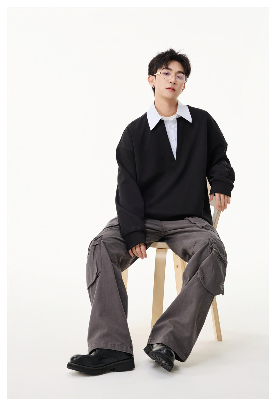 Seated model wearing a black oversized V-neck pullover over a white collared shirt and gray cargo trousers.