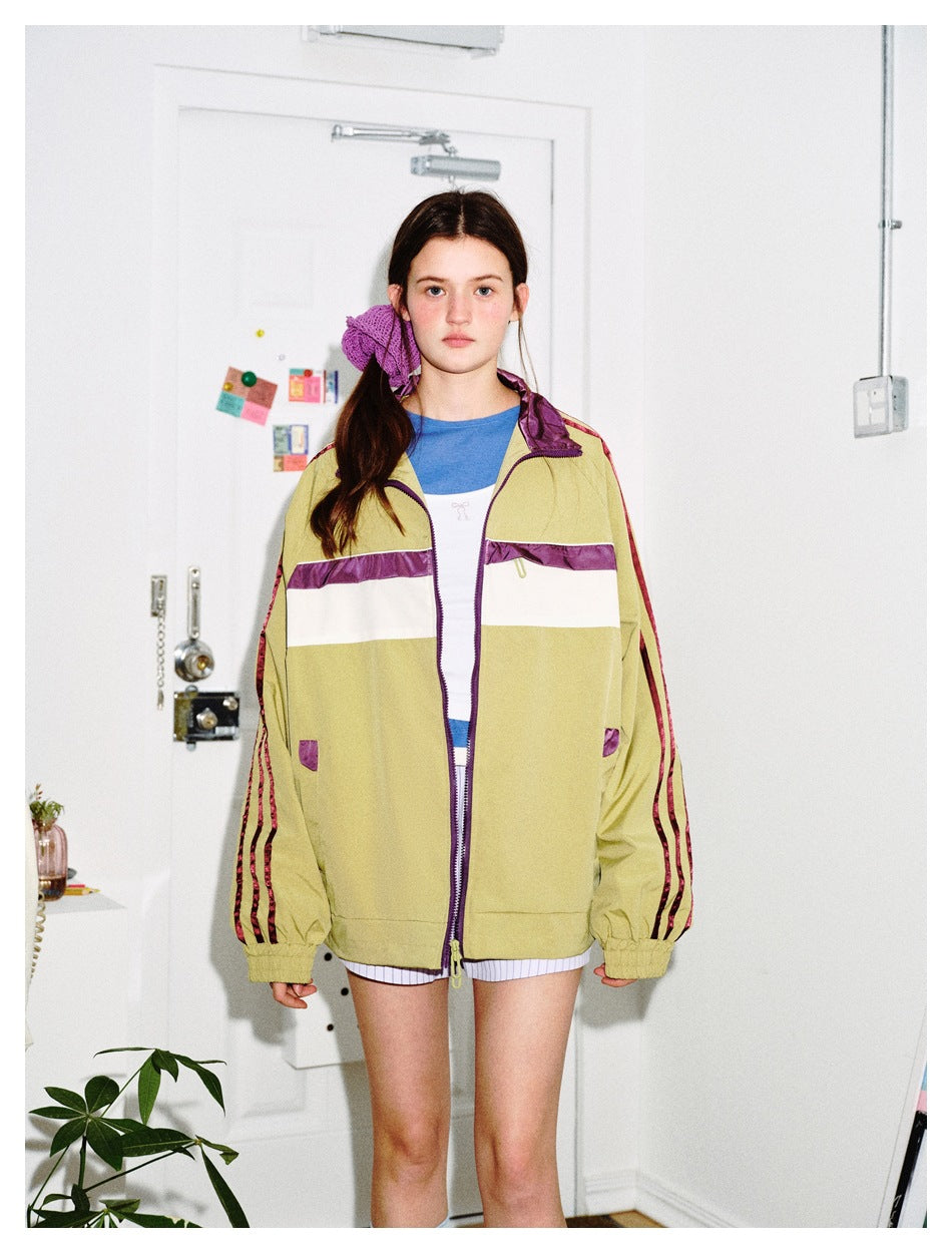 Model wears an open olive track jacket with purple trim, a cream chest stripe and burgundy sleeve stripes.