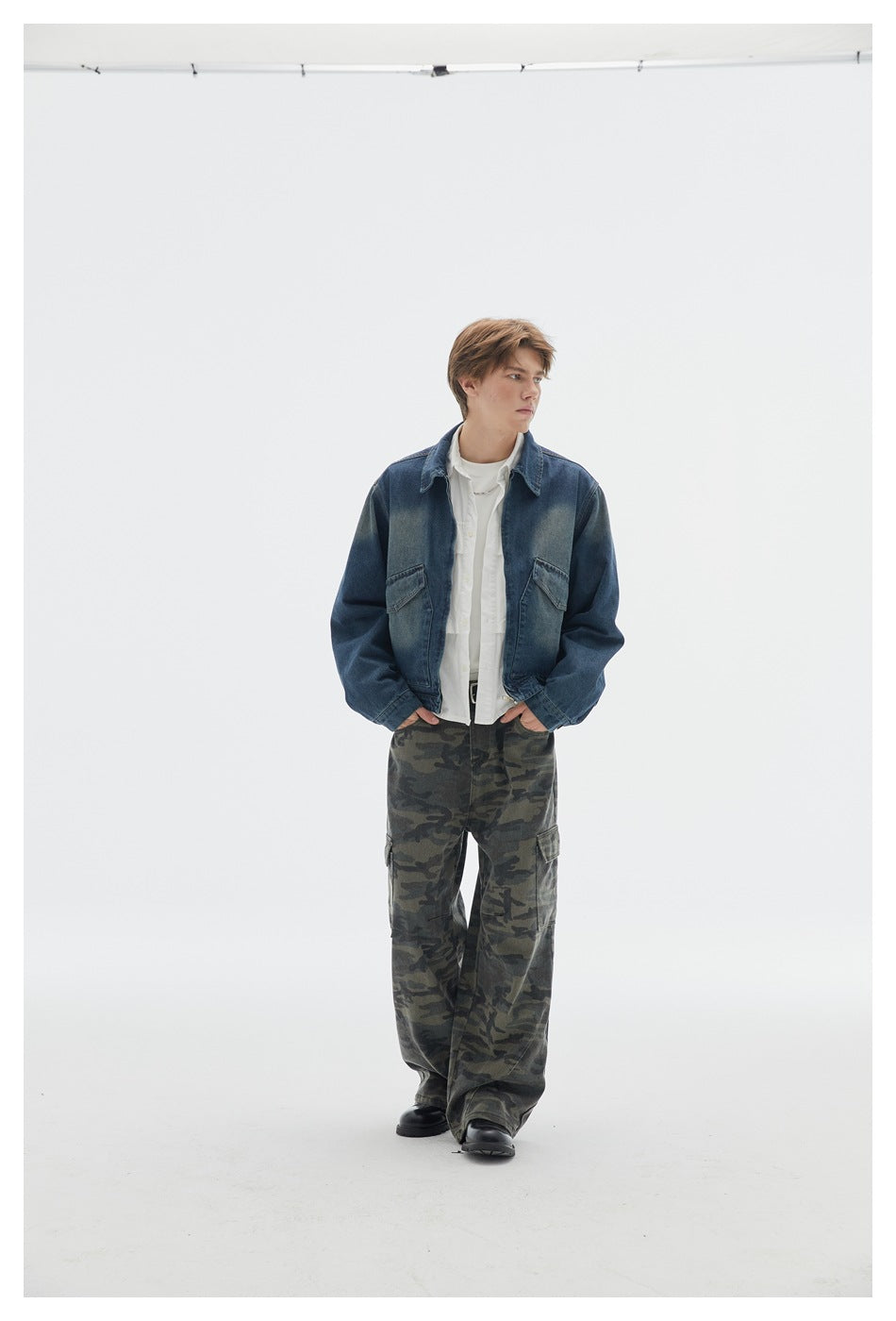 Model wearing a dark blue denim jacket over white layers and loose camouflage cargo pants, hands at the waist.