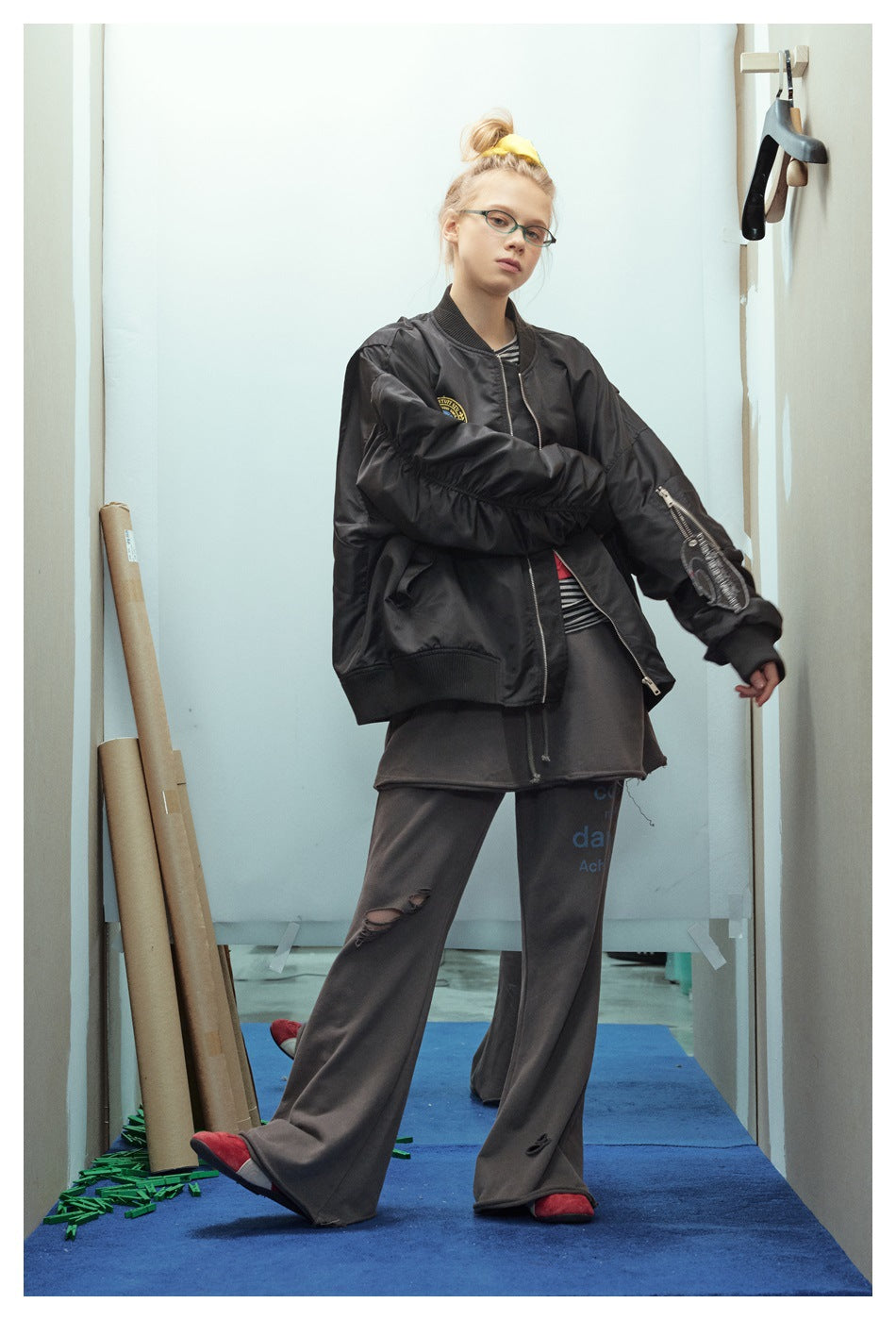 Model wearing a black bomber jacket with ribbed trim, a round chest patch, and a zippered sleeve pocket.