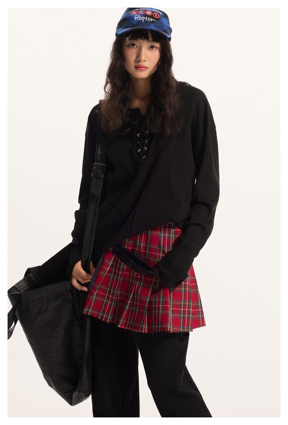 Model wearing a black lace-up top with a red plaid pleated skirt over black trousers.