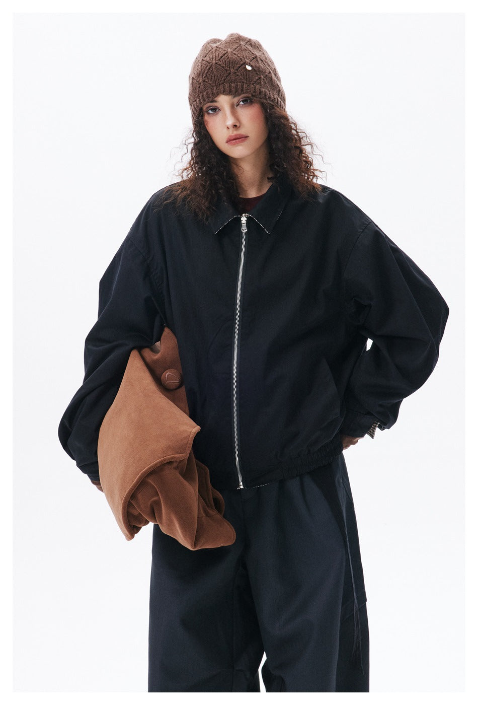Model wearing a black zip-front jacket and brown knit beanie, holding a brown garment against a white studio background.