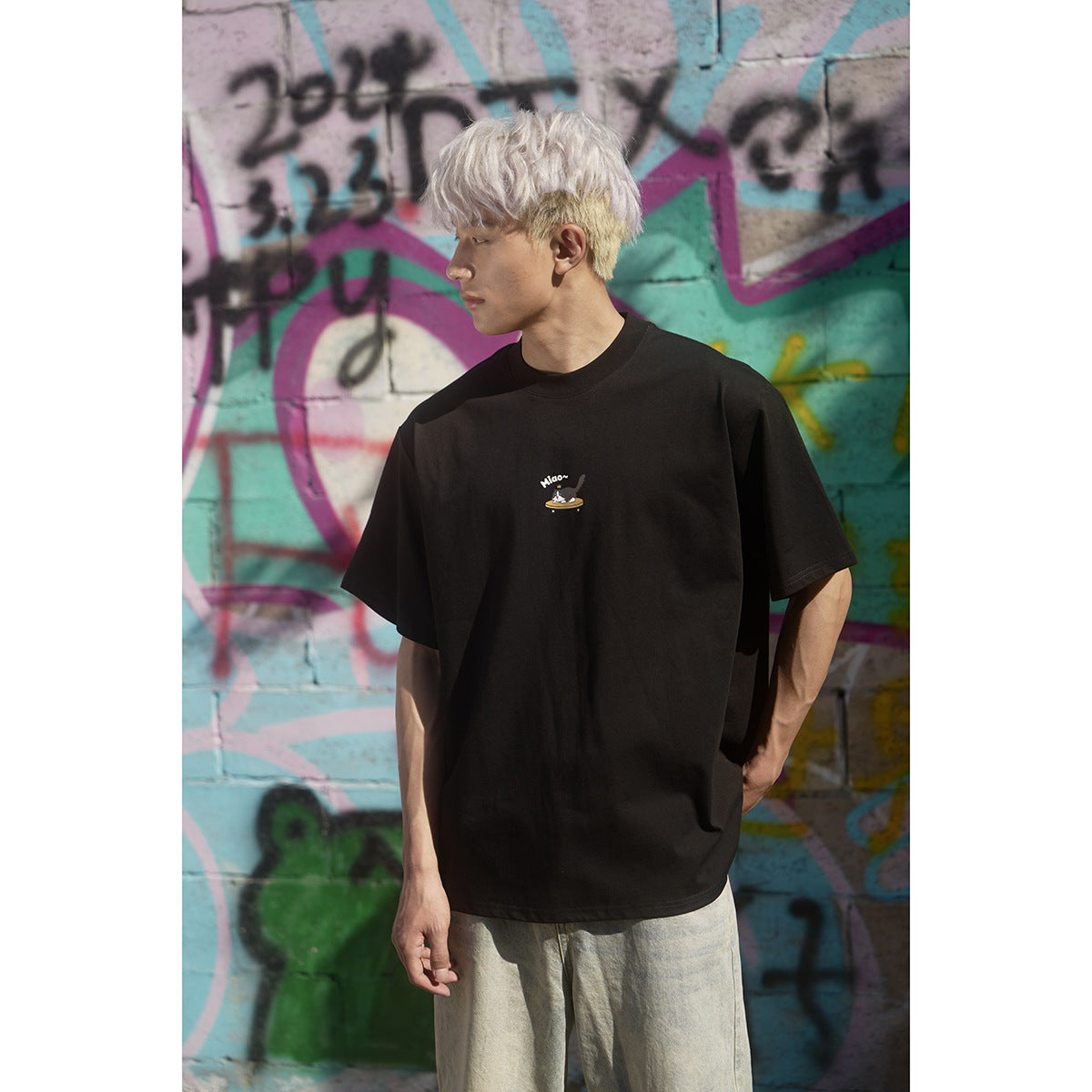 Model wearing an oversized black crew-neck T-shirt with a small illustrated chest graphic, against a colorful graffiti wall.