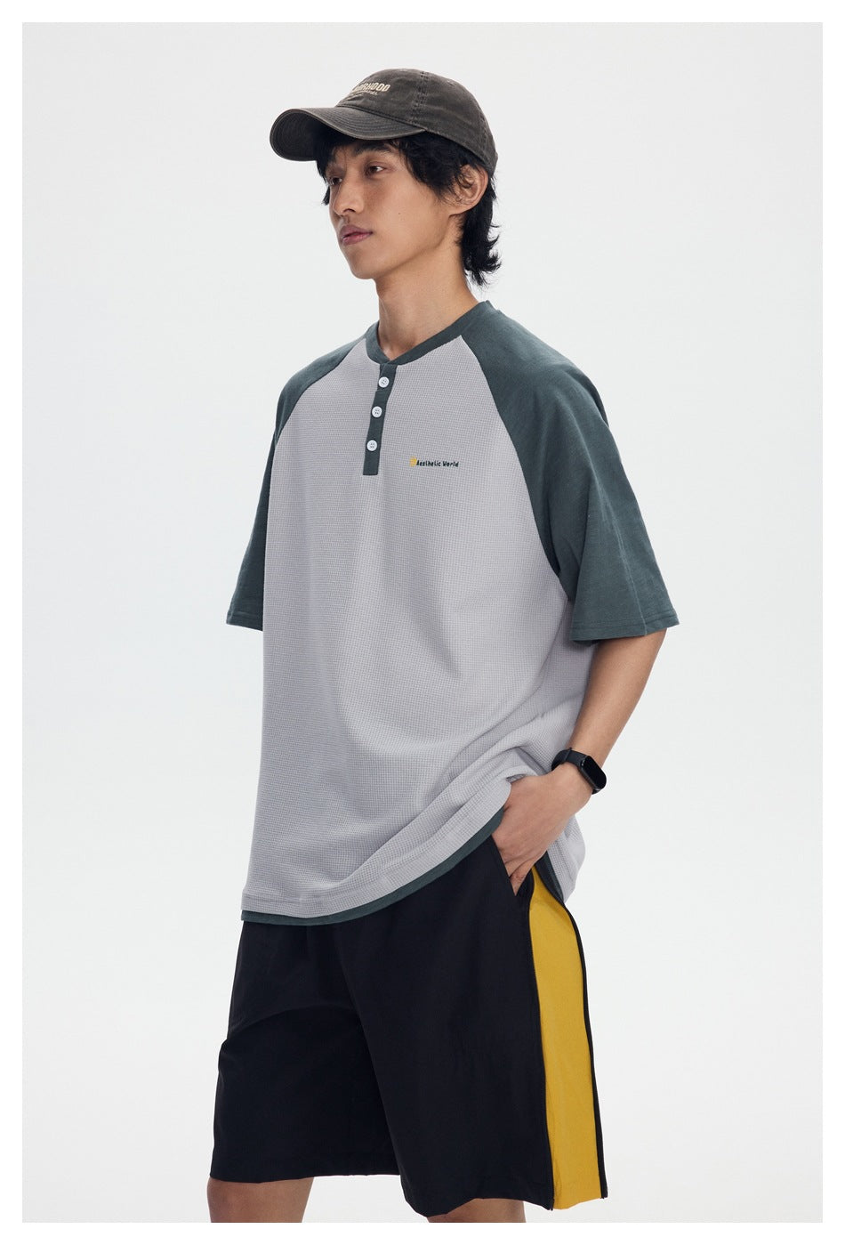 Model wearing a gray raglan henley shirt with dark green sleeves and black shorts with a yellow side panel.