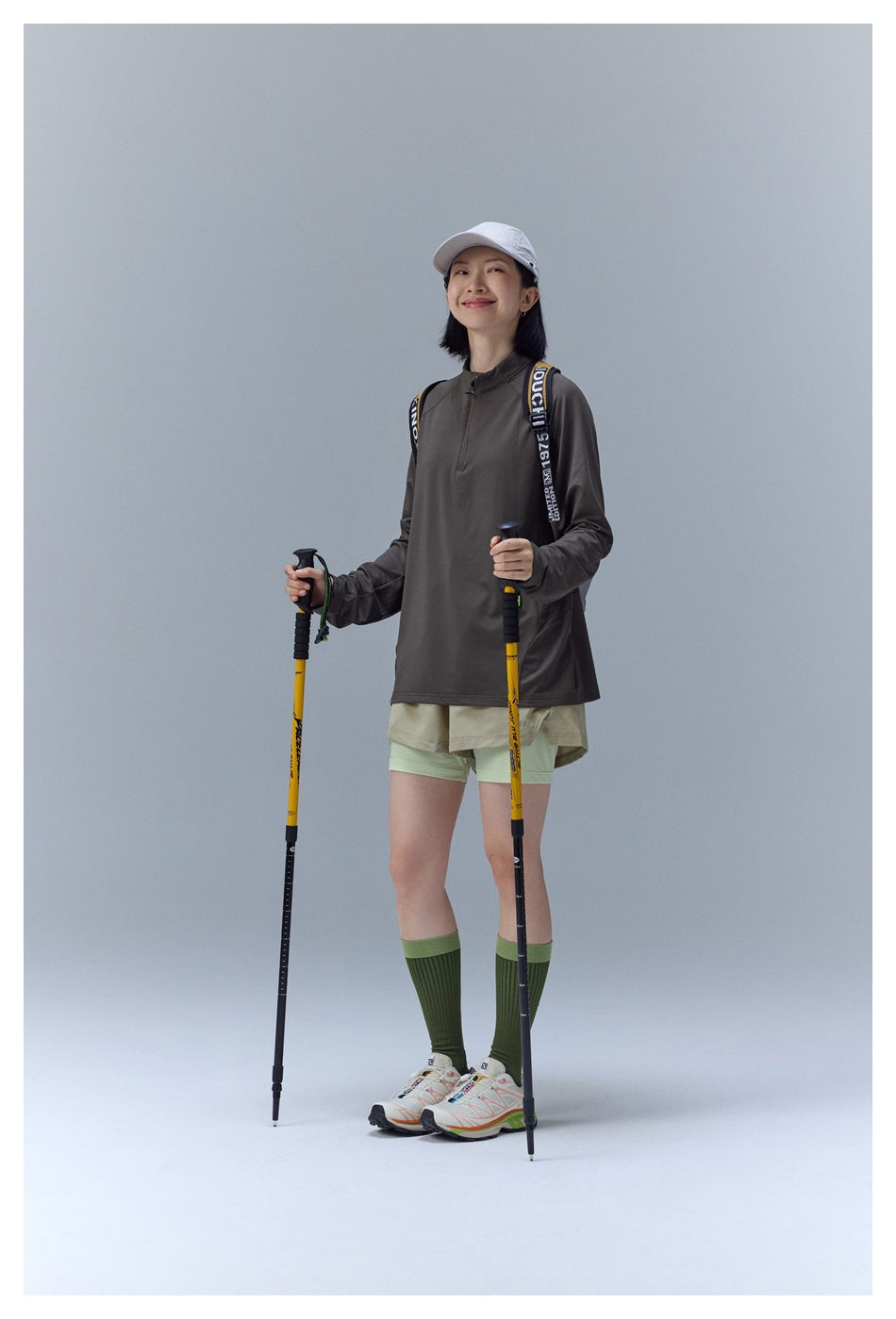 Model wearing a dark taupe quarter-zip top and layered khaki and pale green shorts with hiking poles.