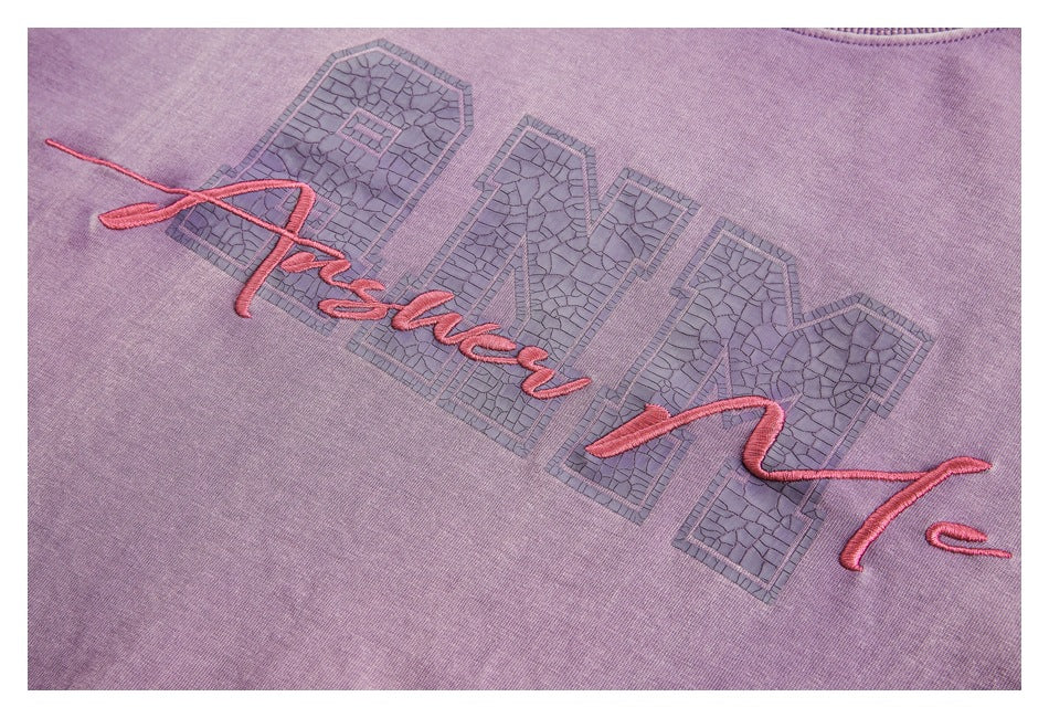 Close-up of a lavender garment with large textured block lettering and pink cursive embroidery.