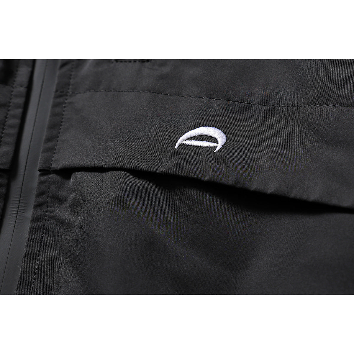 Close-up of a small white embroidered emblem on a black jacket, beside a seam and zipper.