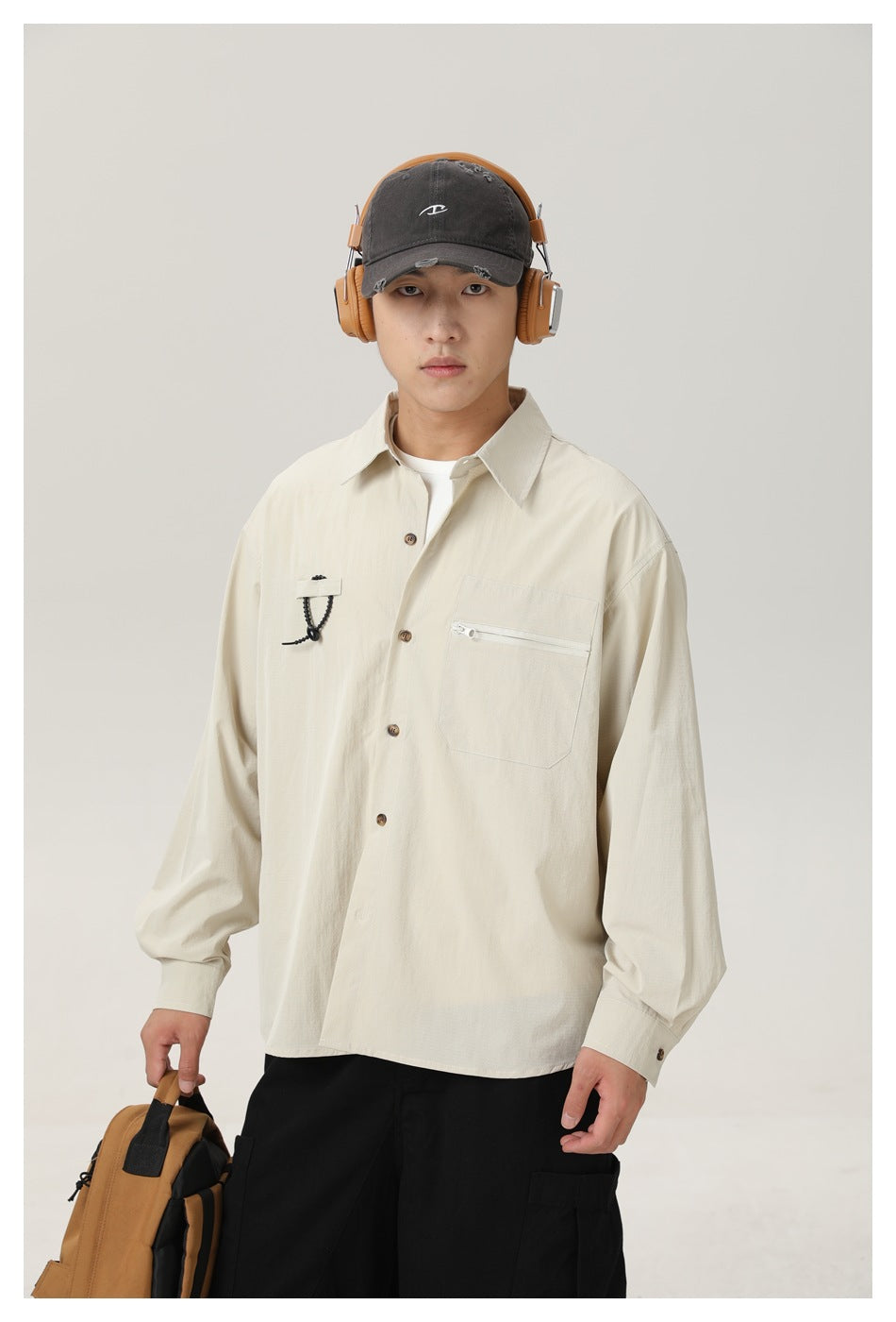 Model wearing an ivory button-up shirt with a zip chest pocket and cord detail, shown with a dark cap and headphones.