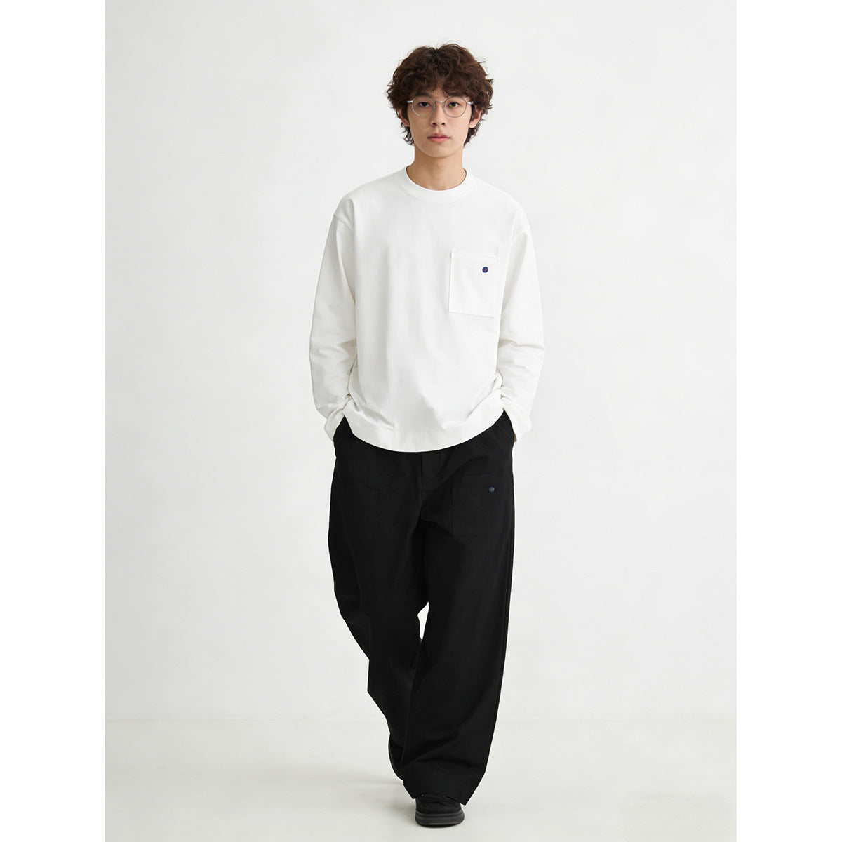 Model wearing a white long-sleeve crewneck shirt with a chest pocket and black trousers.