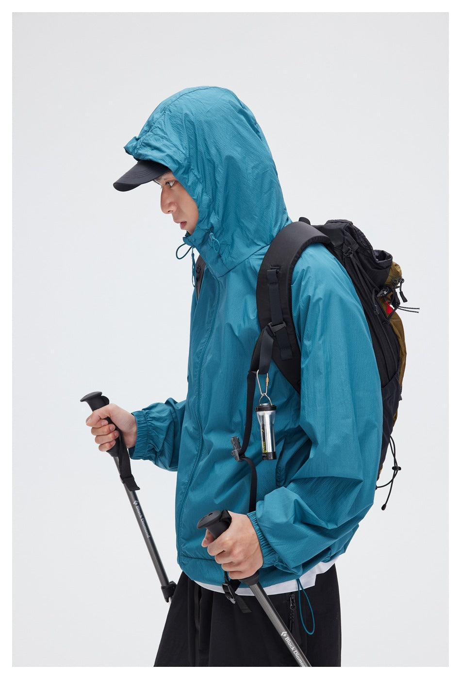 Side view of a model wearing a teal hooded jacket with a black backpack and trekking poles.