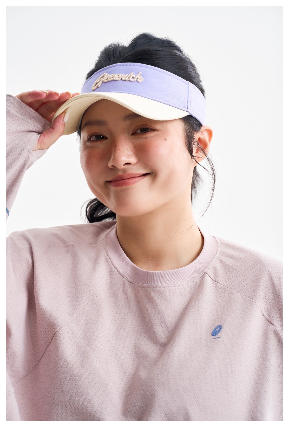 Close-up of a model wearing a lavender visor with embroidered script and a pale pink top with a small blue chest emblem.